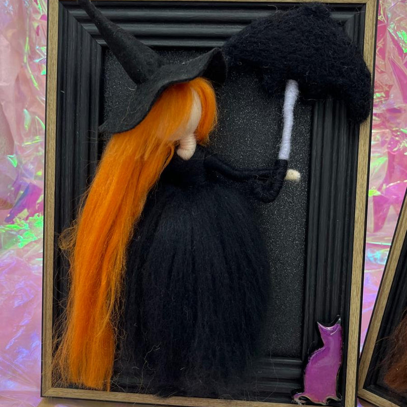 Witch figurine with orange hair and a black hat in a shadow box with a pink and green background.